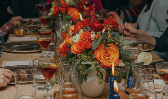 A festive holiday dinner table set with cocktails, wine glasses, and colorful floral centerpieces featuring orange and red roses, surrounded by guests enjoying a meal together.