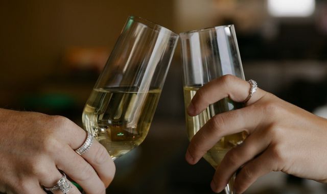 Two hands clink champagne flutes filled with sparkling wine in a softly lit setting.