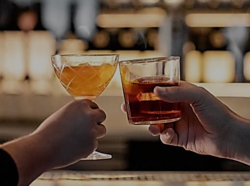 Two hands clink cocktails at a bar, one holding a coupe glass with a golden drink and the other holding a rocks glass with an amber drink, with a softly blurred bar backdrop.
